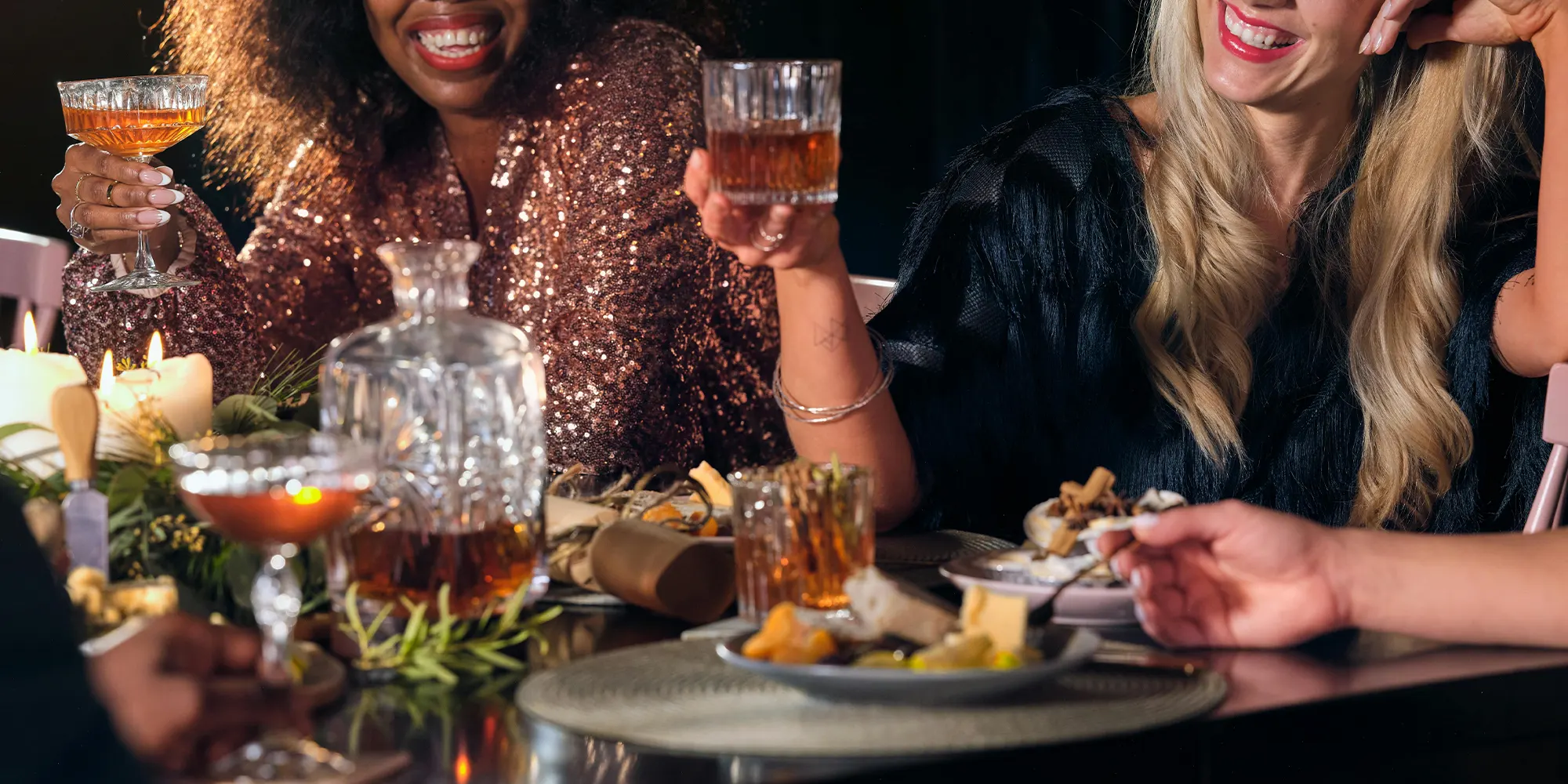 A group of people seated at a table, engaged in conversation, with one person holding a glass of whiskey. The table is set with plates of food, including cheese and vegetables, against a dark background. The people are dressed in formal or semi-formal attire.