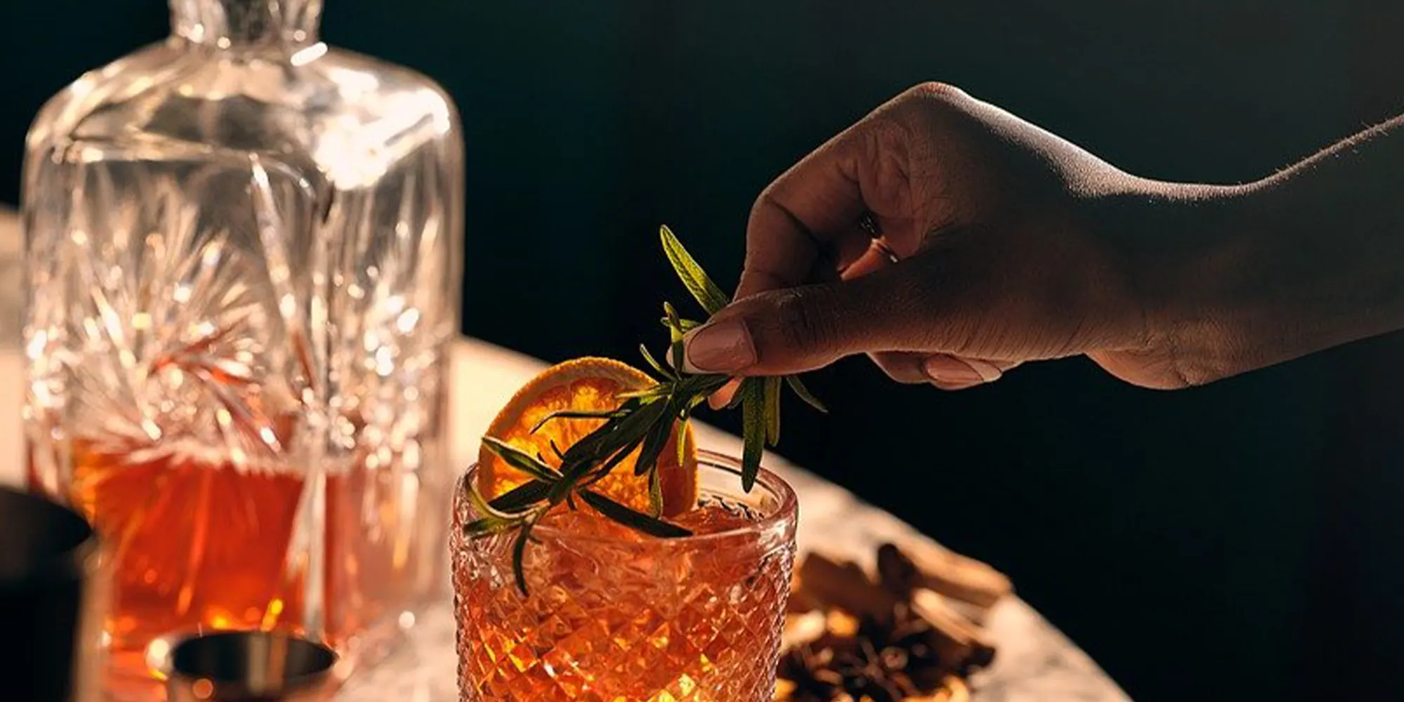 Hand placing a rosemary sprig into an orange cocktail with ice and an orange slice, enhancing the bar's sleek, modern aesthetic.