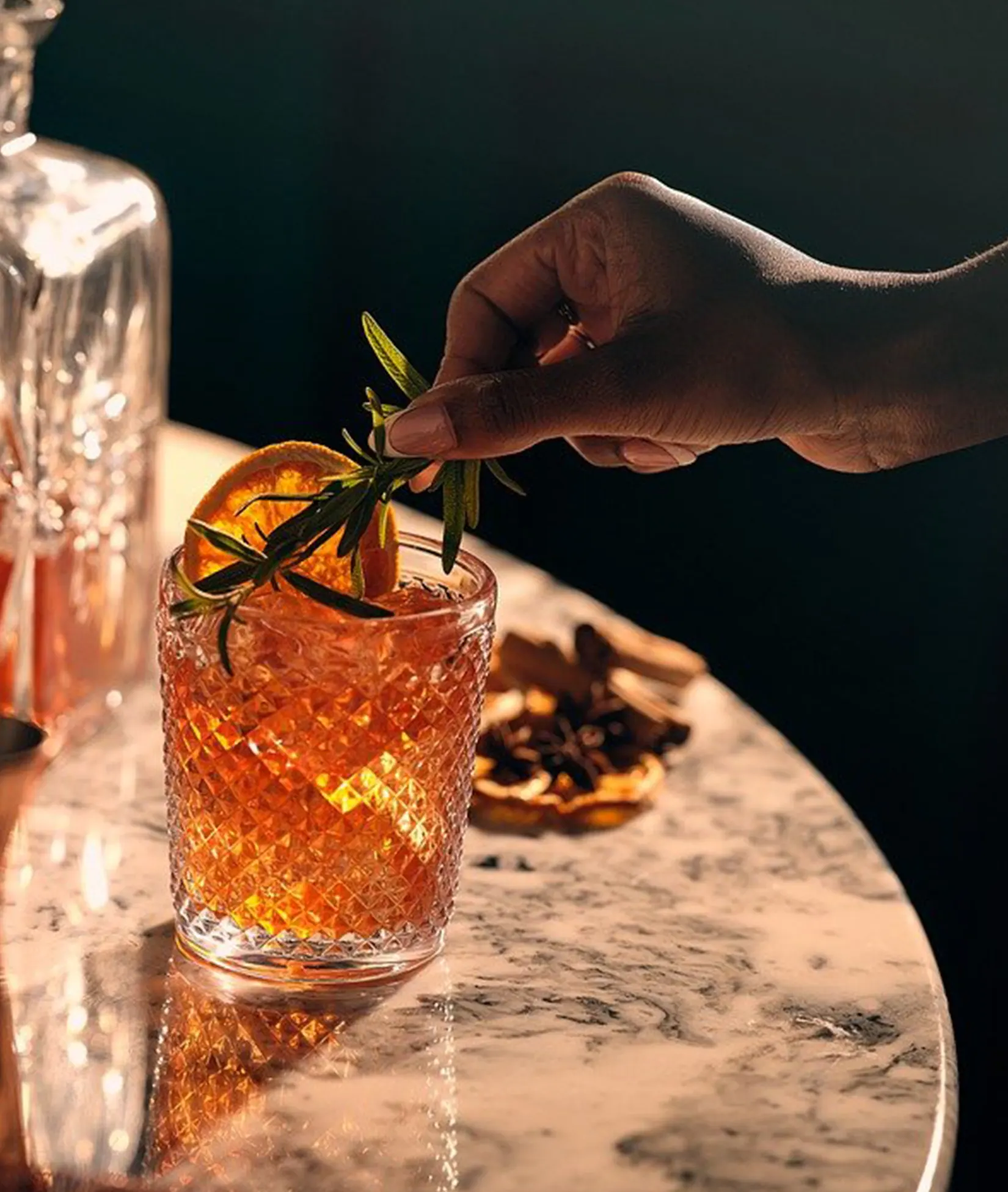 Hand placing a rosemary sprig into an orange cocktail with ice and an orange slice, enhancing the bar's sleek, modern aesthetic.